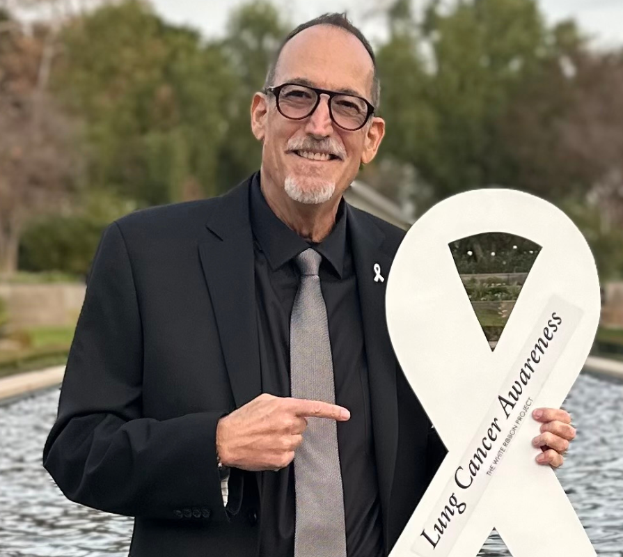 Jeff Stibelman, looking sharp in a black shirt and tie, pointing at the Lung Cancer Awareness Ribbon he's holding